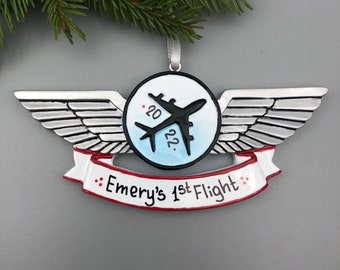 Könnte beinhalten: Silberner Flugzeugflügel-Anhänger mit einem blauen und weißen Kreis, der ein schwarzes Flugzeug und die Daten 2022 und 20 zeigt. Ein rotes und weißes Banner lautet "Emery's 1st Flight".