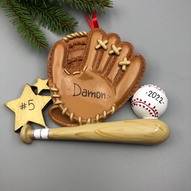 May include: A brown baseball glove with a white baseball and a wooden bat. The glove has the name "Damon" written on it. A gold star with the number "5" is also included. The baseball has the year "2022" written on it.