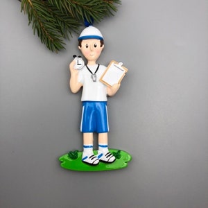 May include: A white and blue Christmas ornament depicting a person wearing a white shirt, blue shorts, a white hat, and holding a stopwatch and a clipboard.