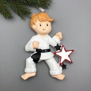 May include: A white karate uniform ornament with a black belt, a red and white star, and black stars. The ornament features a boy with blonde hair in a karate pose.