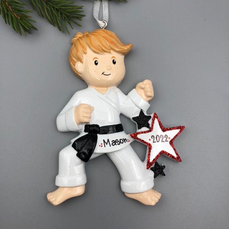 May include: White personalized Christmas ornament of a boy in a karate uniform with a black belt. The ornament has a red star with black glitter and the text "2022" on it. The boy's name "Mason" is written on the belt.