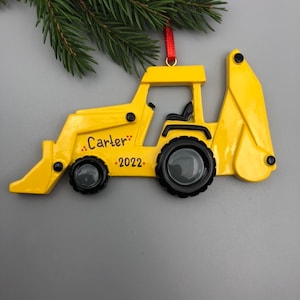 May include: Yellow toy backhoe ornament with black wheels and a red ribbon. The ornament is personalized with the name "Carter" and the year "2022".