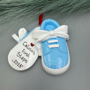 May include: Blue baby shoe ornament with a white bow and a white tag that says "Calvin's First Steps 2023" with a red heart.