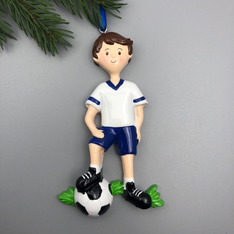 May include: A white and blue soccer player ornament with brown hair and a black and white soccer ball. The ornament is standing on green grass.