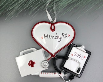 May include: White and red heart ornament with the text "Mindy, RN" and the year "2023". The ornament also features a stethoscope, a first aid kit, and a clipboard.