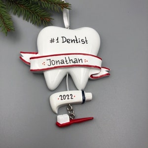 May include: White tooth-shaped Christmas ornament with a red ribbon that says "#1 Dentist Jonathan" and a red and white toothbrush and toothpaste hanging from it. The ornament also has the year "2022" on it.