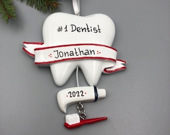 May include: White tooth-shaped Christmas ornament with a red ribbon that says "#1 Dentist Jonathan" and a red and white toothbrush and toothpaste hanging from it. The ornament also has the year "2022" on it.