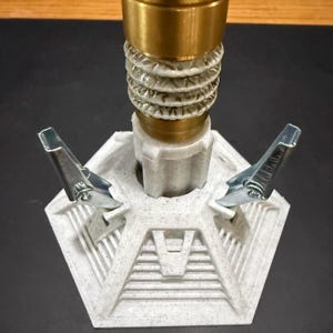 May include: A close-up of a light gray and gold-colored object. The object has a hexagonal base with two silver clips attached. The top portion is gold, and the middle section is light gray with a textured design.