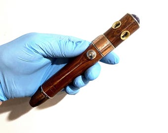 May include: A handcrafted wooden pen with a dark brown finish, accented with copper and silver metal bands. The pen features a purple gemstone at the top and is held in a gloved hand. The pen's design is both elegant and functional.