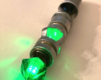 May include: A silver and green, light-up, handheld prop resembling a futuristic device. The device has a green crystal at the end and a series of silver rings.