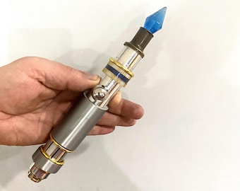 May include: A silver and gold-colored sonic screwdriver with a blue crystal tip. The screwdriver has a textured, metallic finish and a small, round button on the side.