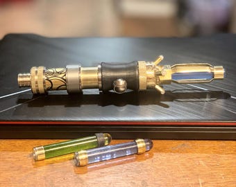 May include: A gold-colored, steampunk-style device with a black rubber grip and a clear glass tube. The device has a series of gears and levers. Two small, clear glass tubes with colored liquid are also pictured.