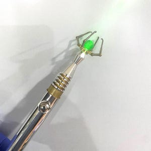 May include: A silver and brass tool with a green glass sphere at the end. The tool has a button on the side and a claw-like mechanism at the top.