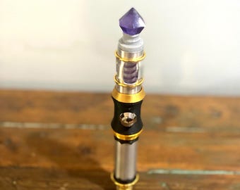 May include: A silver and black vaporizer with gold accents and a purple crystal top. The device has a cylindrical design with a visible coil and a button. The vaporizer is displayed on a wooden surface.
