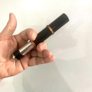 Könnte beinhalten: Ein schwarzer und silberner Teleskop-Stift mit einem Kupferband. Der Stift wird in einer Hand gehalten.