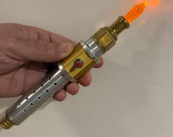 May include: A cylindrical prop device featuring a silver handle with perforated holes, a gold-colored midsection with a small red circular button, and a gold-toned tip emitting a bright orange light from a textured, translucent, flame-shaped emitter.