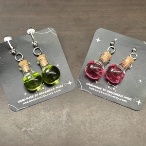 May include: A pair of earrings featuring miniature glass potion bottles with cork stoppers. One pair contains green liquid, the other pink. The earrings are displayed on black cards with the text "HANDMADE BY NEKONEKO MINI nekonekomini.etsy.com".