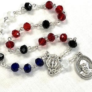 May include: A rosary with faceted beads in red, black, white, and blue, strung on a silver chain. The rosary includes a silver pendant with an image of a saint and a smaller pendant with a religious emblem.
