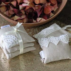 May include: Four beige linen sachets with white lace trim and a white satin ribbon. The sachets are stacked on top of each other. One sachet has a white lace trim and a white satin ribbon. The sachets are on a gold-coloured lace doily.