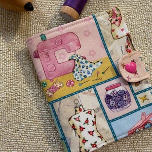 May include: A patchwork sewing kit featuring a pink sewing machine, dress form, and button jar design. The kit has a pink heart-shaped button closure. A purple reel of thread and a wooden hedgehog are also in the image.