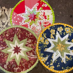 May include: Three circular quilted trivets with starburst patterns. One has a red and white design, another features a floral print, and the third has a blue floral pattern. Each trivet has a contrasting border.