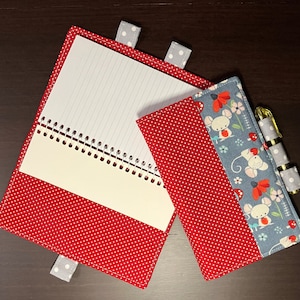 May include: A red and white polka dot notebook with a spiral binding and a smaller red and white polka dot pouch attached. The pouch has a blue and white fabric lining with a pattern of mice and flowers.