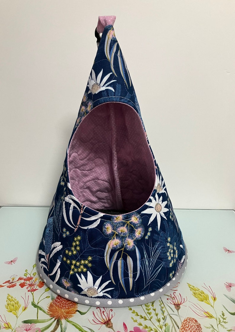 K&ouml;nnte beinhalten: Ein blaues und wei&szlig;es Blumenmuster-H&auml;ngebett f&uuml;r Haustiere mit rosafarbenem gestepptem Innenraum. Das Bett hat einen grauen und wei&szlig;en Tupfenrand.