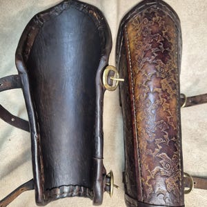May include: A pair of dark brown leather arm guards. One guard is plain, while the other features an embossed floral pattern. Both have brass buckles and leather straps for securing them to the arm. The leather has a worn, antique appearance.