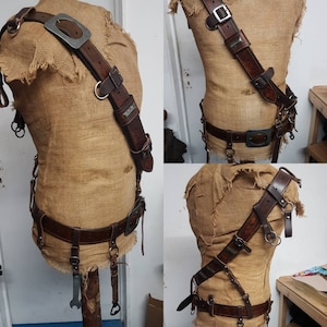 May include: A brown leather harness with metal buckles and rings, displayed on a burlap-covered mannequin. The harness includes a shoulder strap, belt, and various attachments, showcasing a rugged, handcrafted design.