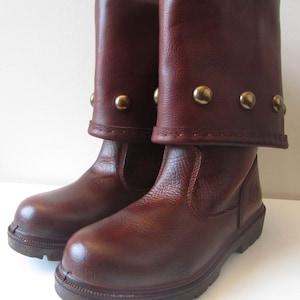 May include: A pair of brown leather boots with a fold-down cuff and gold metal accents. The boots have a chunky sole and a rounded toe.