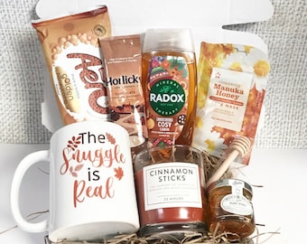 May include: A gift box containing self-care products. It features a mug with the text "The Snuggle is Real", a candle, honey, a face mask, Radox shower gel, Horlicks chocolate, and Aer honeycomb.