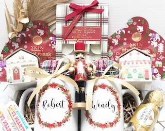 May include: A gift box filled with holiday-themed items. Includes two white mugs with names, a nutcracker, skincare products, candy, and chocolates. The mugs are decorated with a wreath design. The box is tied with a gold ribbon.
