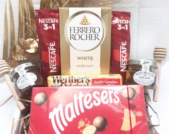 May include: A gift basket filled with various treats. It includes a box of Maltesers, a box of Ferrero Rocher White Hazelnut chocolates, Werther's Butter Candies, Nescafe 3-in-1 packets, jars of honey, and honey dippers. Gold decorative elements are also included.