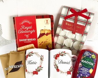 May include: A gift box containing a variety of items. Includes a box of shortbread fingers, two mugs with floral wreaths and names, tea lights, coffee packets, and a bottle of shower gel. The box is white with a red ribbon.