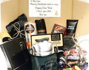 May include: A gift box filled with various items, including a mug that says "Collect Moments Not Things", a can of coffee, a bottle of body spray, a package of hand warmers, a bar of soap, a black washcloth, a bottle of cologne, and a bag of candy. The box is decorated with brown paper and a handwritten note that says "To My Cam, Merry Christmas and a very Happy New Year. I love you lots. Bec xx"