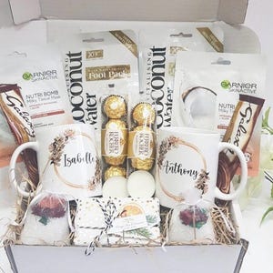 May include: A white gift basket filled with self-care items, including two mugs with the names "Isabelle" and "Anthony", a chocolate bar, face masks, bath bombs, and other bath and body products.