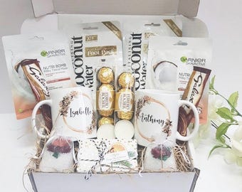 May include: A white gift basket filled with self-care items, including two mugs with the names "Isabelle" and "Anthony", a chocolate bar, face masks, bath bombs, and other bath and body products.
