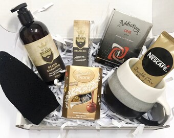 May include: A gift box filled with a variety of items, including a black and white mug, a bottle of beard shampoo, a bottle of beard oil, a box of chocolates, a bottle of cologne, and a packet of instant coffee. The box is filled with white shredded paper.