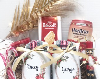 May include: A gift box filled with festive items, including two personalized mugs with floral designs and names, a box of Biscoff white chocolate, and a jar of Horlicks pudding. Candy canes and a nutcracker are also included.
