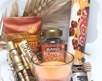 May include: A gift box filled with various treats. It includes a bag of honey roasted nuts, a jar of coffee, a candle, a jar of honey, a roll of Rolo candies, a Werther's Original candy, and a gold nutcracker. The candle is labeled "Chocolate Orange".