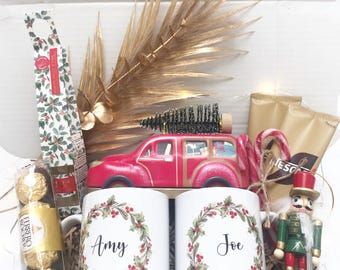 May include: A gift box filled with holiday-themed items. Includes two white mugs with names, a red wooden car, a nutcracker, candy canes, chocolates, and decorative gold accents. The mugs are decorated with a wreath design.