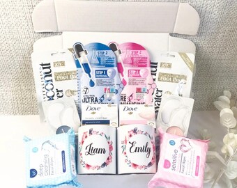 May include: A white gift box filled with beauty products. Includes facial masks, foot pads, Dove beauty cream bars, facial wipes, and two personalized mugs with floral designs and the names "Liam" and "Emily."