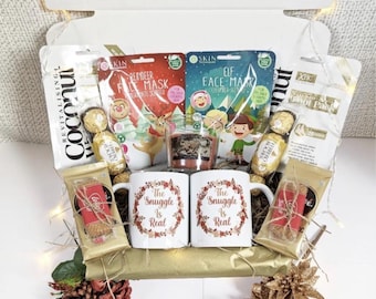 May include: A gift box filled with holiday-themed items. Includes face masks, mugs with "The Snuggle Is Real" text, chocolates, a candle, and other treats. The box is white with a gold base and decorative accents.