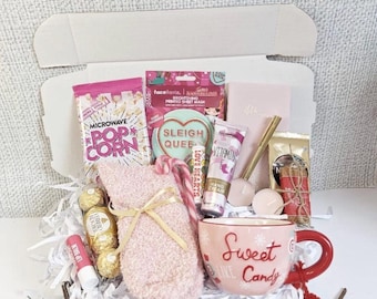 May include: A gift box filled with self-care items. Includes a pink mug with "Sweet Like Candy" text, popcorn, a face mask, lip balm, hand cream, a fuzzy sock, and chocolates. The box is decorated with festive accents.