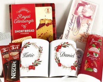 May include: A gift basket with a red and plaid shortbread box, two white mugs with floral wreaths and names, a chocolate bar, shower gel, and Nescafe coffee packets. The mugs are personalized.