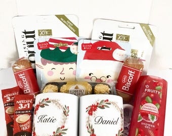 May include: A gift box filled with various items, including face masks, coffee packets, Biscoff treats, Ferrero Rocher chocolates, and personalized mugs with floral wreaths and names. A red shower gel bottle is also visible.