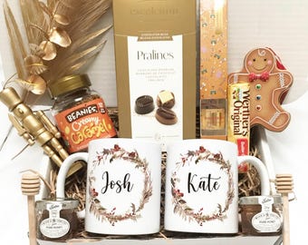 May include: A gift basket filled with treats, including two white mugs with floral wreaths and names "Josh" and "Kate", a box of chocolates, coffee, honey, and a gingerbread man. The basket is decorated with gold accents.