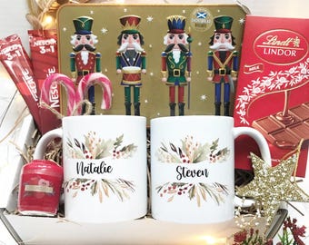 May include: A gift box with Christmas-themed items. Includes two white mugs with floral designs and names, a tin of nutcracker biscuits, a bar of Lindt chocolate, coffee packets, a red candle, and candy canes. Decorative lights and ornaments complete the festive arrangement.