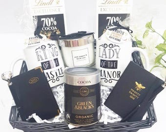 May include: A wicker basket filled with a gift set featuring a black and white mug with the text "King of the Castle", a black and white mug with the text "Lady of the Manor", a black journal with gold text "Twenty Twenty One", a black and gold cocoa powder tin, a white candle with a black label, and two chocolate bars with gold text.