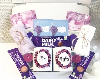 May include: A gift box filled with self-care items. Includes two mugs with names, Cadbury Dairy Milk chocolate, lavender-scented face masks, small towels, candles, and instant hot chocolate packets. The box is white, and the items are arranged inside.